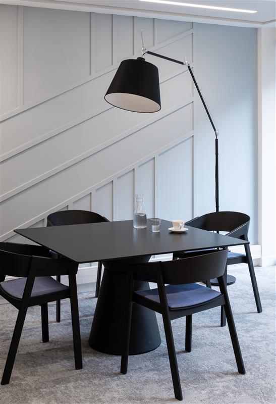 Petite salle de réunion avec table et chaise noir, une grande lampe amène la lumière au dessus de la table, mur blanc et sol gris clair
