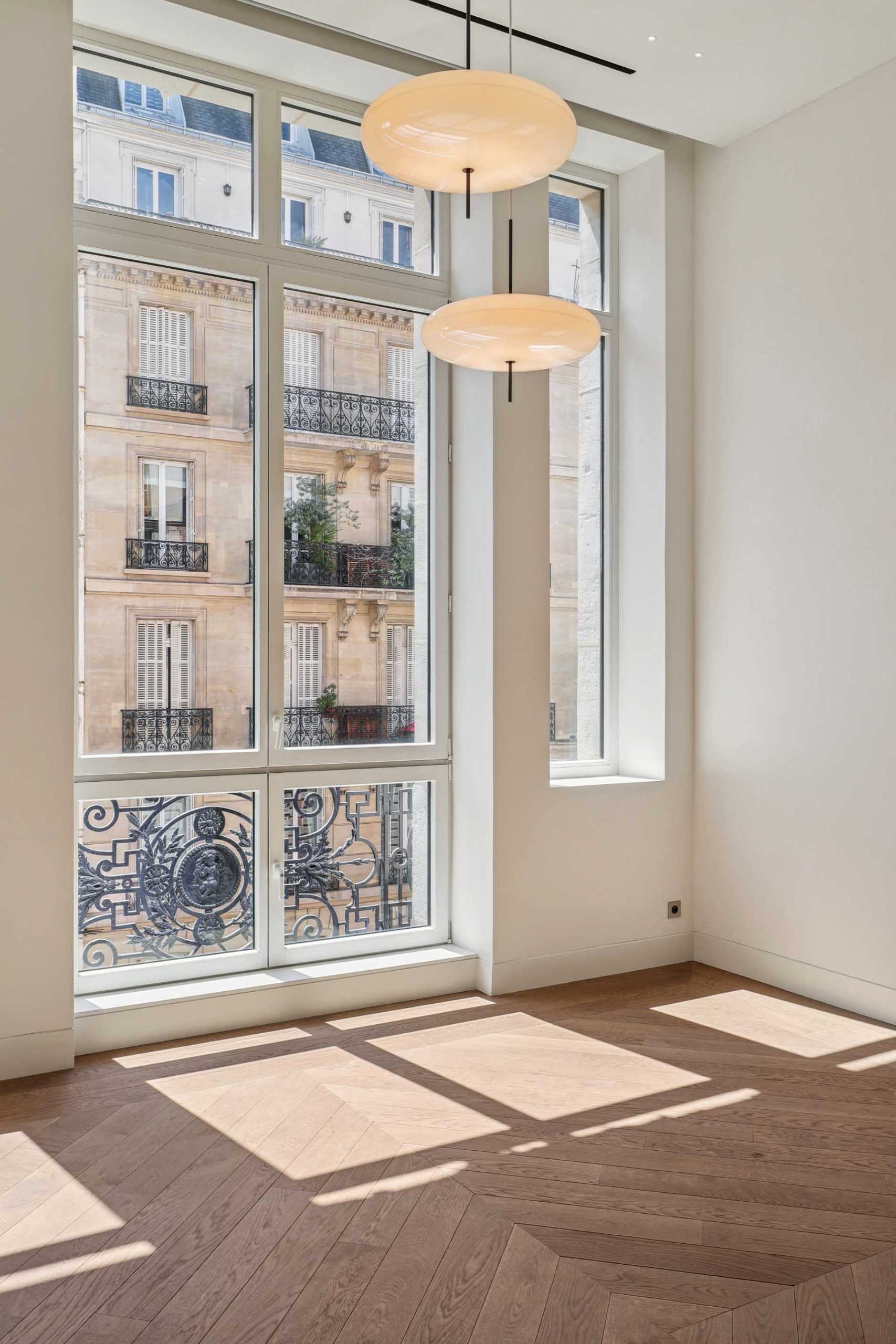 Grande porte fenêtre amenant sur un balcon, pièce aux murs blancs et luminaires suspendus