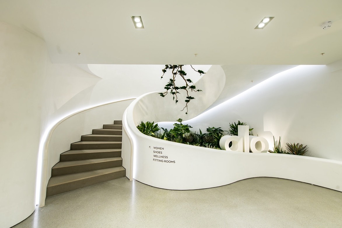 Escalier en béton blanc, avec lumière sur les rambardes, stickage en bas pour indiquer le type de produits à l'étage et Logo en volume au milieu de plantes décoratives.