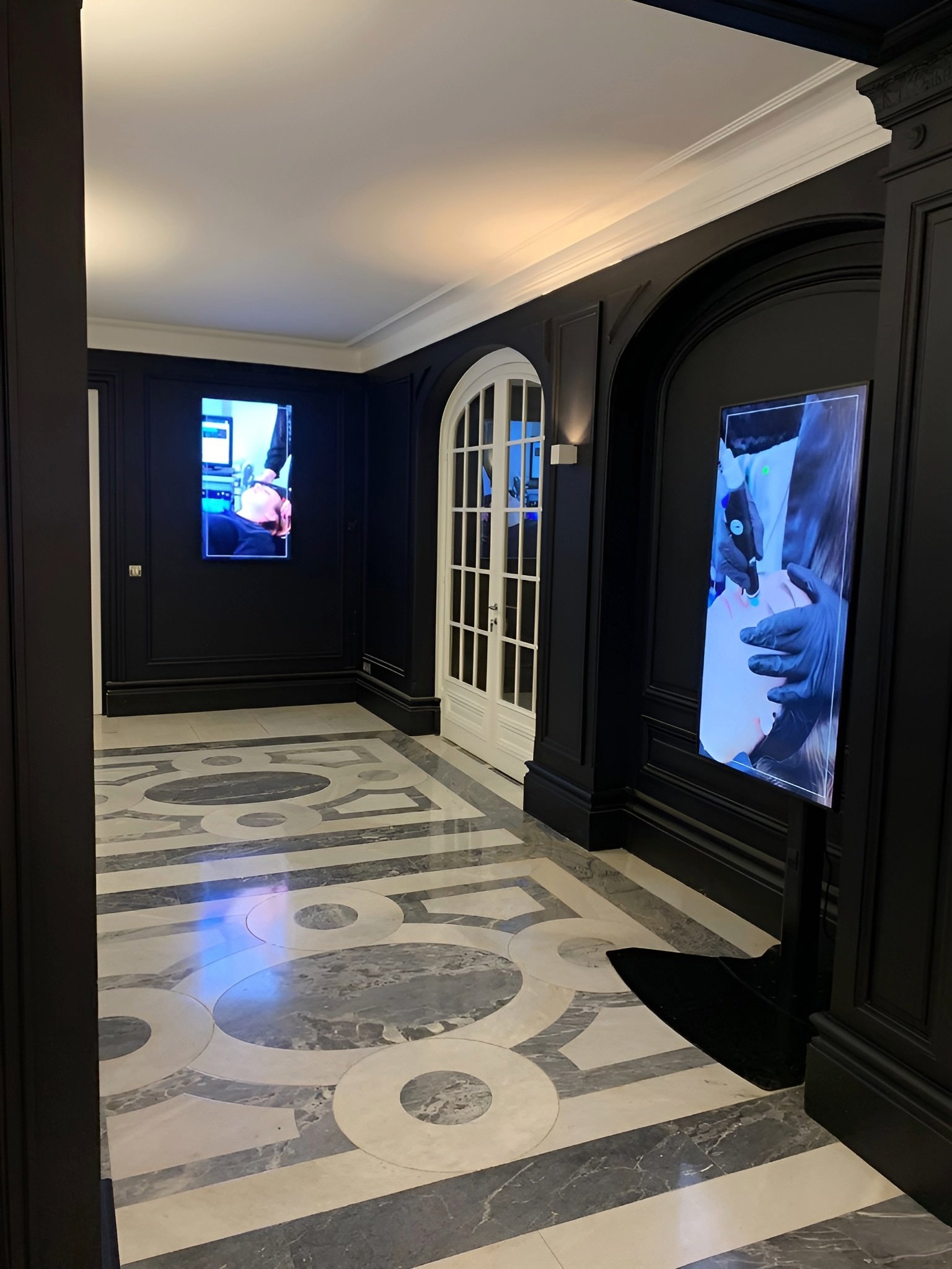 Espace avec mur peint en noirs, plafond blanc, sol en carrelage blanc et gris clair, écran vidéo au mur
