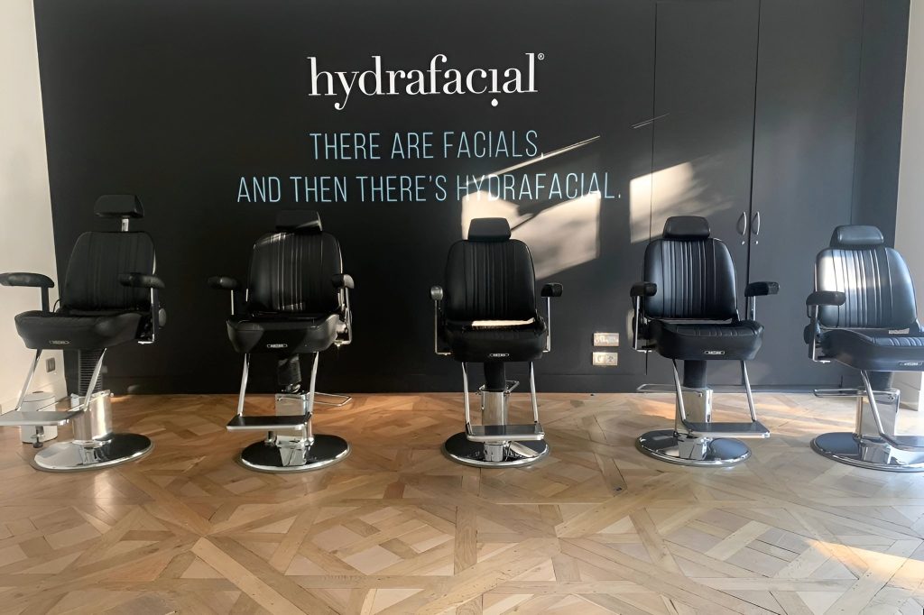 Espace avec plusieurs fauteuils de barbier en cuir noir, mur du fond peint en noir avec le logo de la marque en blanc