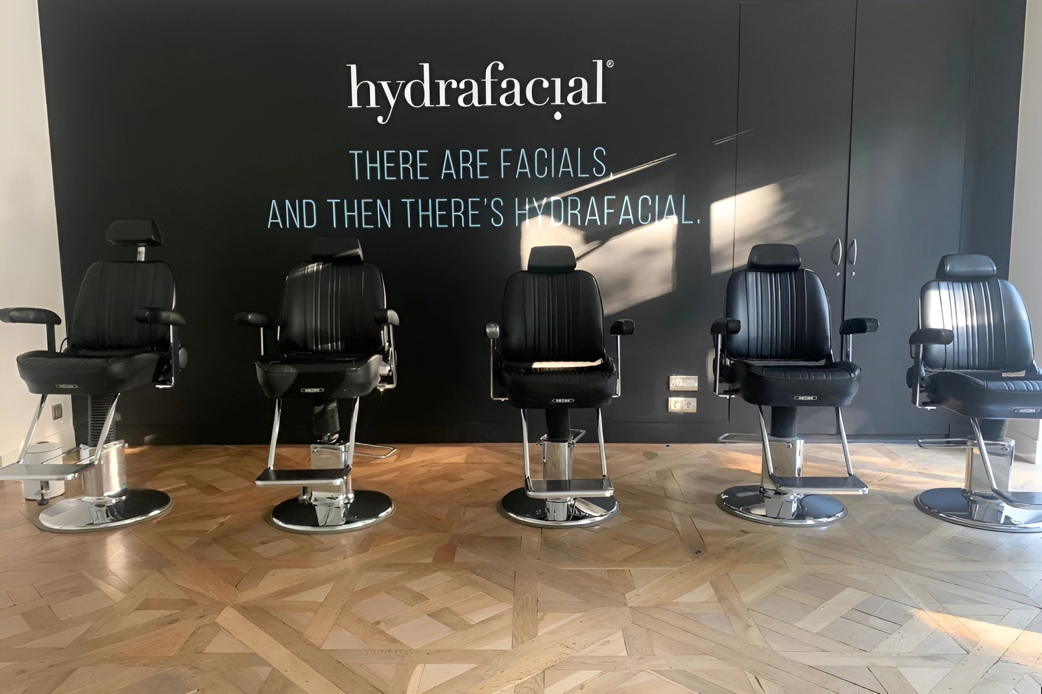 Espace avec plusieurs fauteuils de barbier en cuir noir, mur du fond peint en noir avec le logo de la marque en blanc