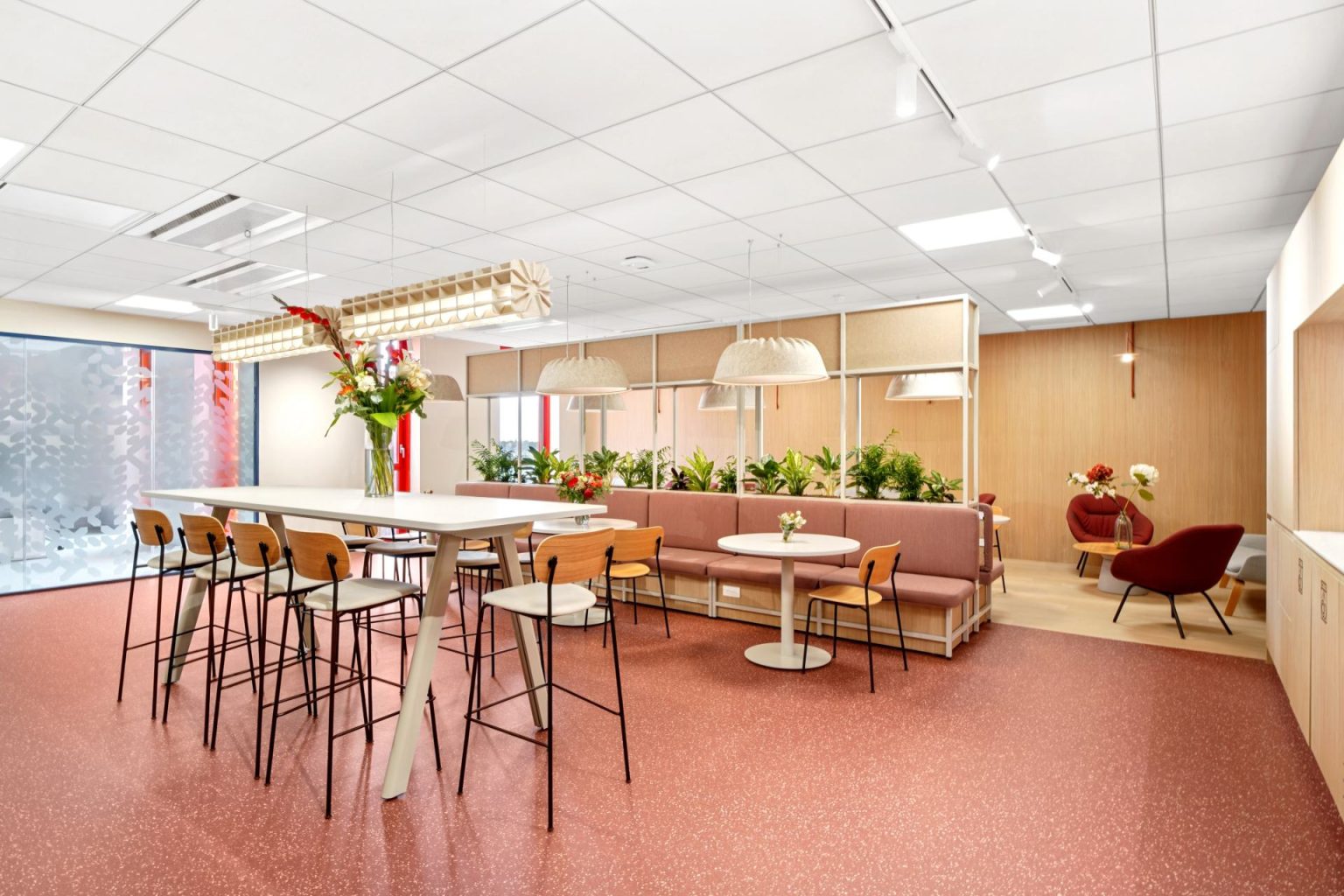 Espacio de coworking, suelo rojo, sillas y mesas altas, separador con banco con cojín de color ocre, acabado en madera clara.